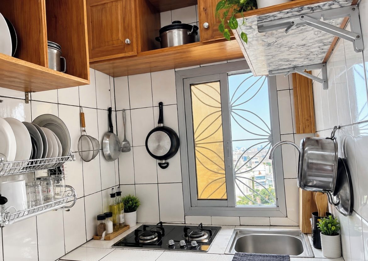 A kitchen with white tiles, wooden cabinets, a gas stove, hanging pots, and a window with a decorative view.