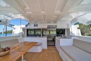 Interior de barco com área de jantar, assentos e pequena cozinha.