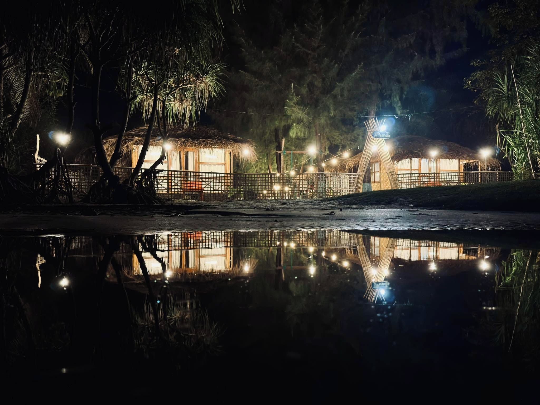 Une scène nocturne avec des huttes aux toits de chaume et des guirlandes lumineuses, dont la lumière se reflète sur la surface calme de l’eau.