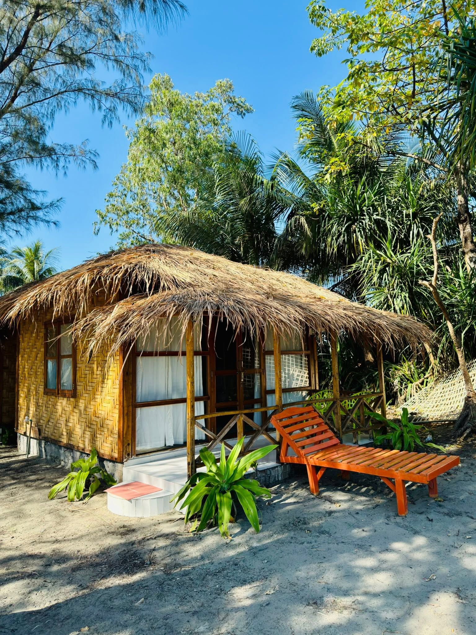 Eine strohgedeckte Hütte mit einer Holzveranda, einem einzelnen Liegestuhl und einem kleinen Gartenbereich mit tropischen Pflanzen.