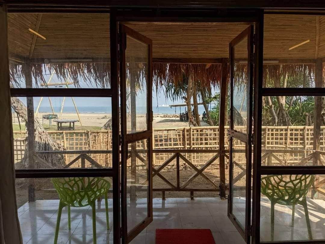 Une vue sur la plage à travers une porte vitrée avec un cadre en bois, un tapis rouge et des chaises vertes.