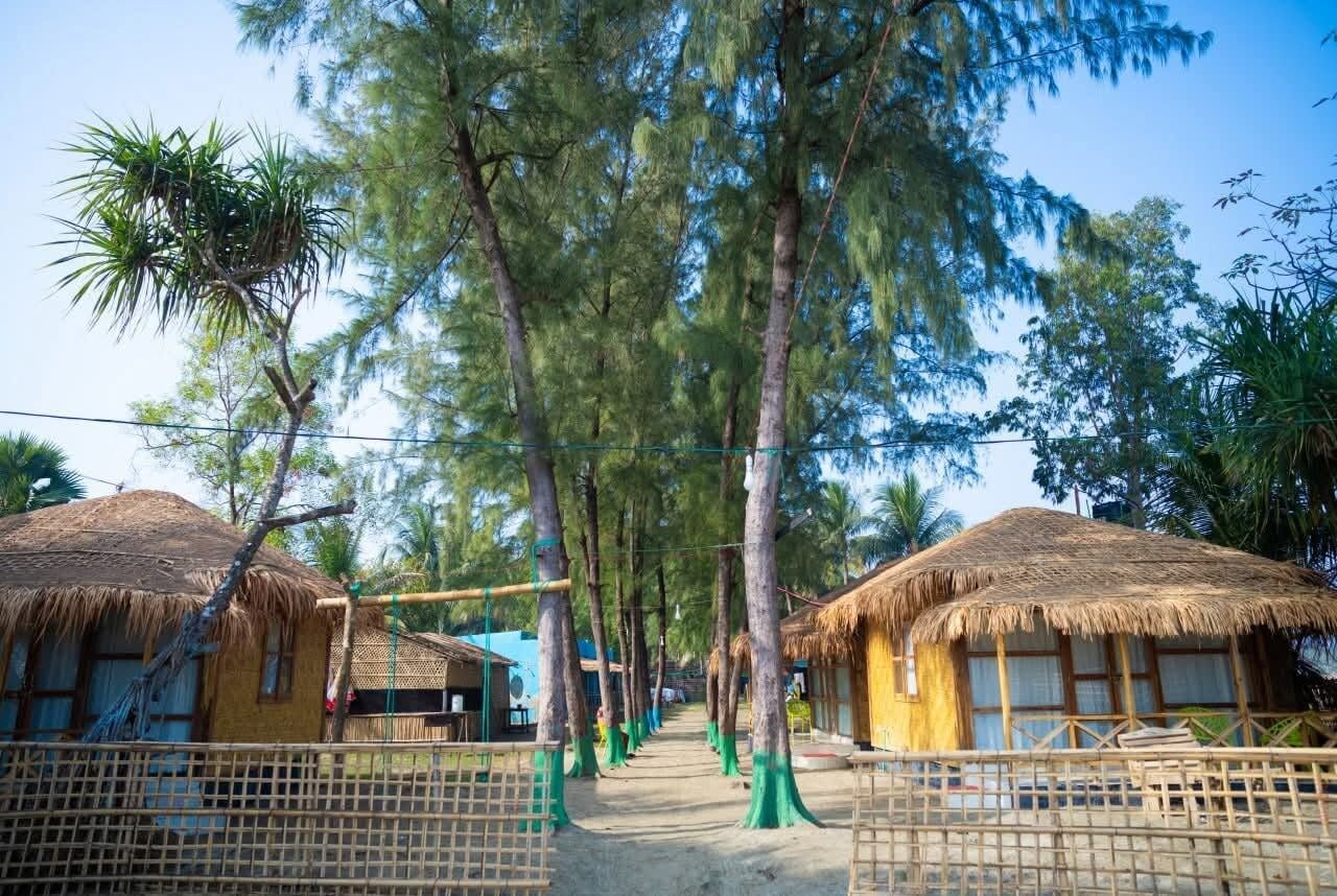 A pathway lined with thatched-roof huts, surrounded by tall trees.