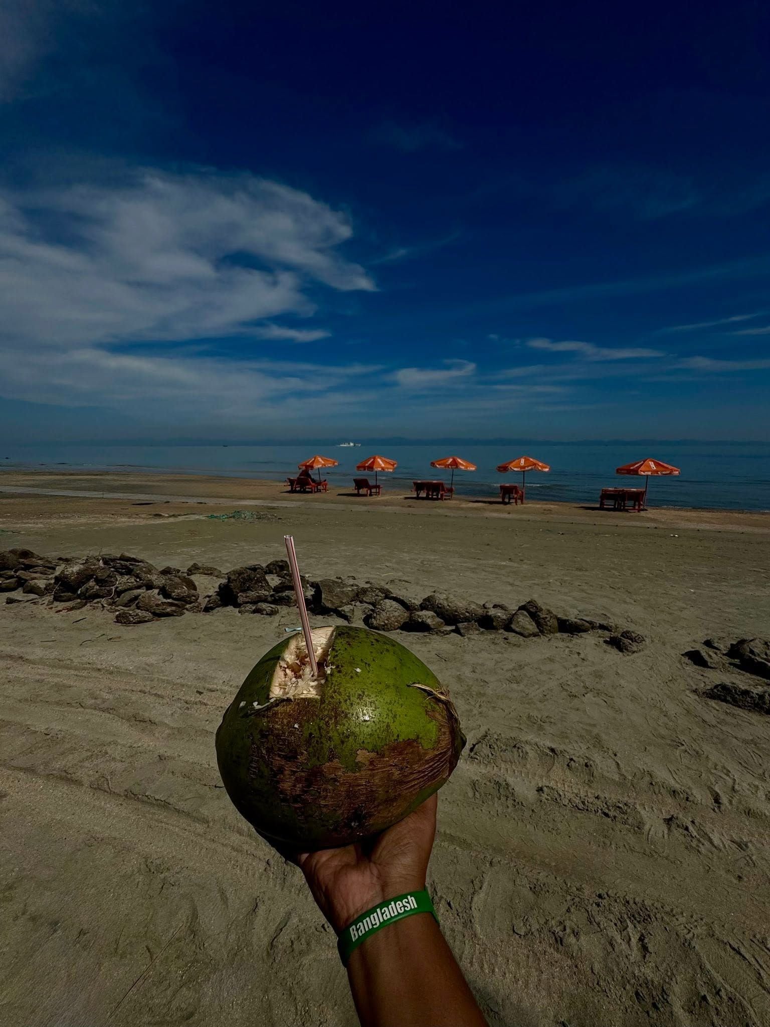 Escena de playa con un coco en la mano, cielo despejado y varias sombrillas de playa al fondo.