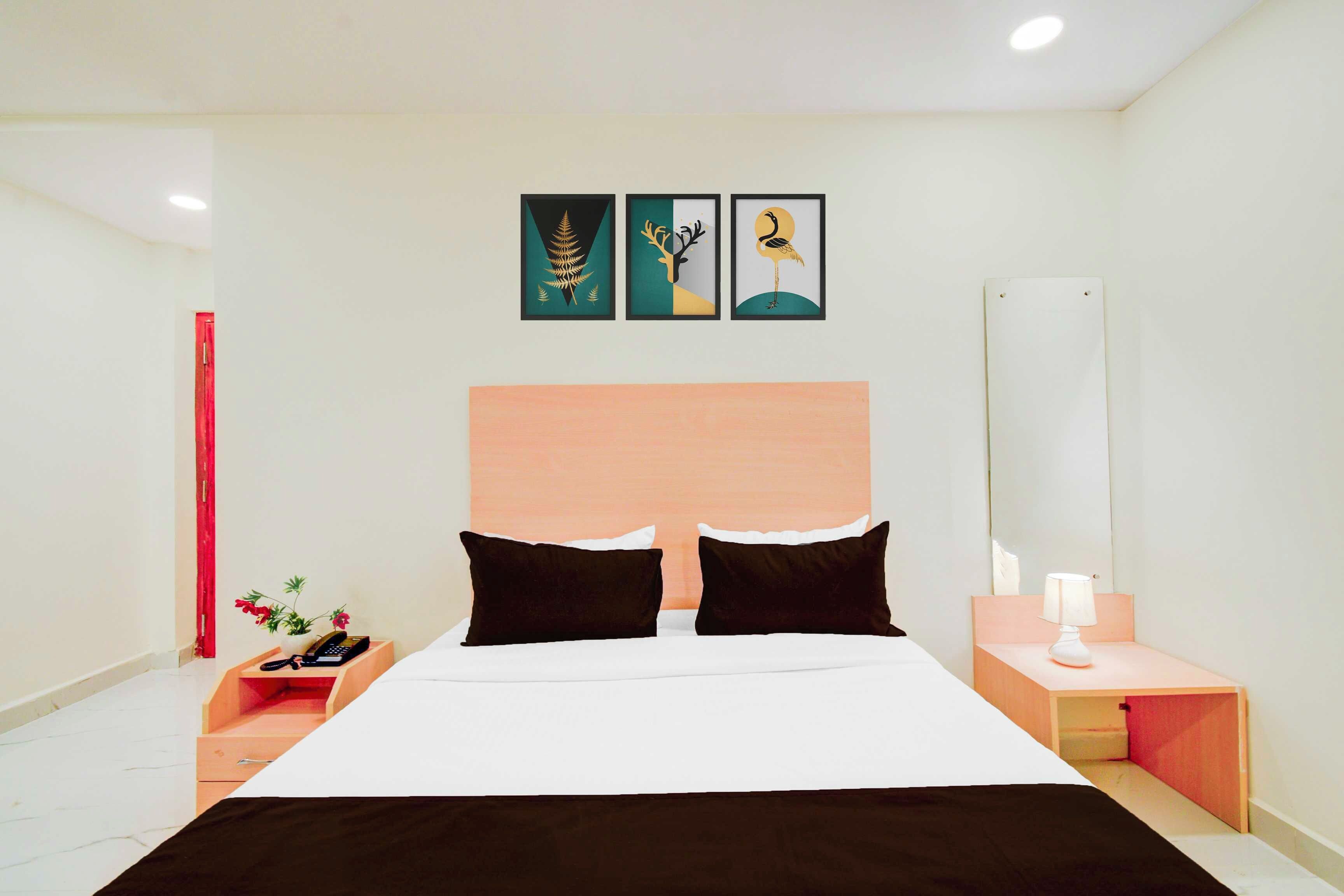 Una habitación de hotel con una cama, dos almohadas negras, una mesita de noche con una lámpara, una pequeña mesa con un teléfono y flores, y tres obras de arte enmarcadas en la pared.