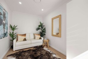 Ein weißes Ledersofa mit zwei Kissen, ein kleiner Holztisch, ein Spiegel, eine große Kübelpflanze und ein flauschiger Teppich.