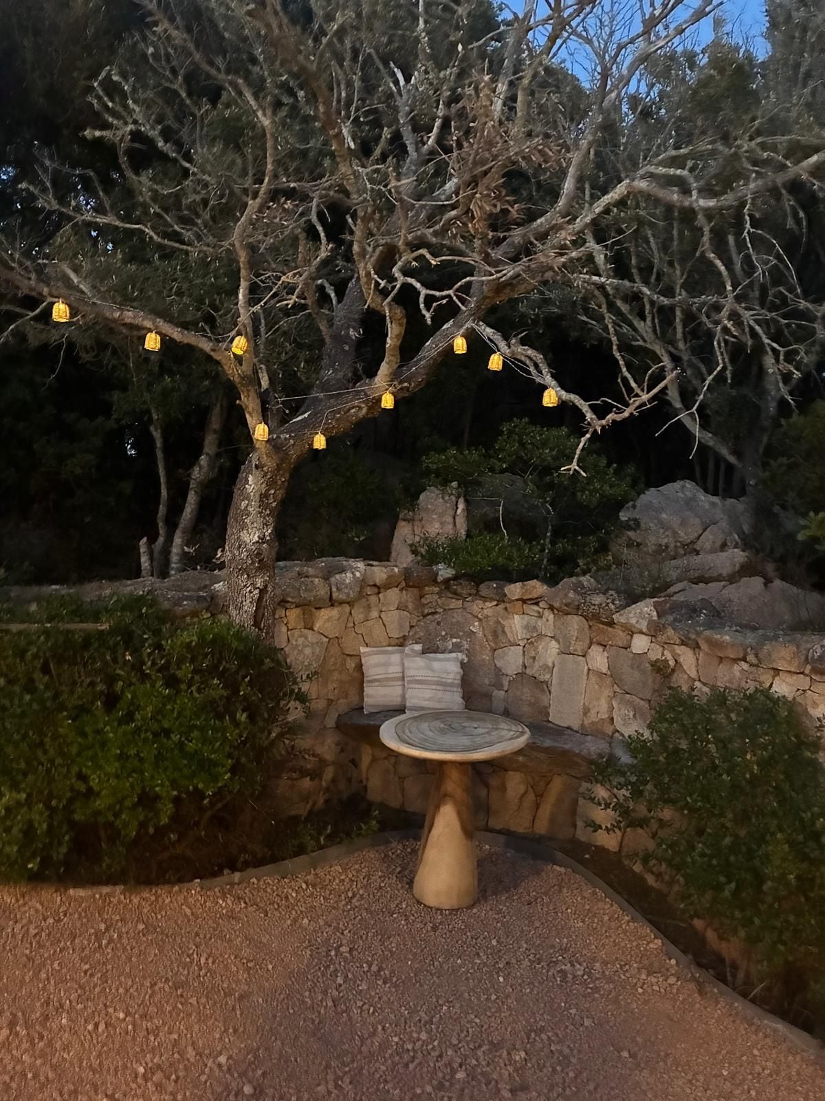 A stone bench with cushions, surrounded by a gravel area, under a tree with hanging yellow lights.