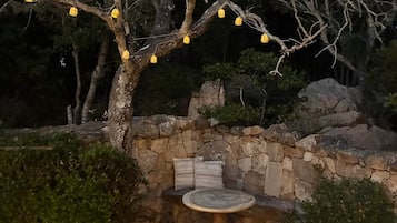 A stone bench with cushions, surrounded by a gravel area, under a tree with hanging yellow lights.