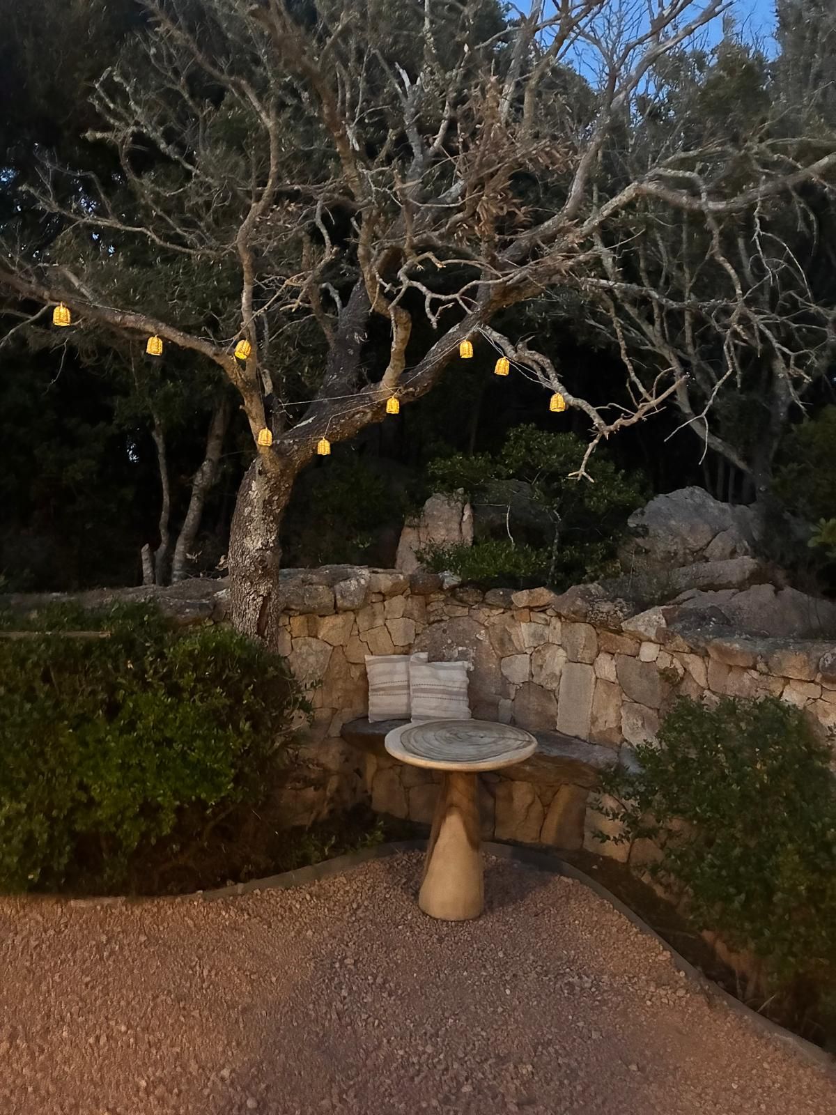 Un banco de piedra con cojines, rodeado por un área de grava, bajo un árbol con luces amarillas colgantes.