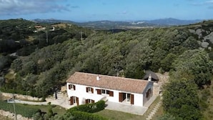 A single-story building with a tiled roof, surrounded by dense greenery and hills in the background. - Stazzu La Vigna di Maria (Sardegna)