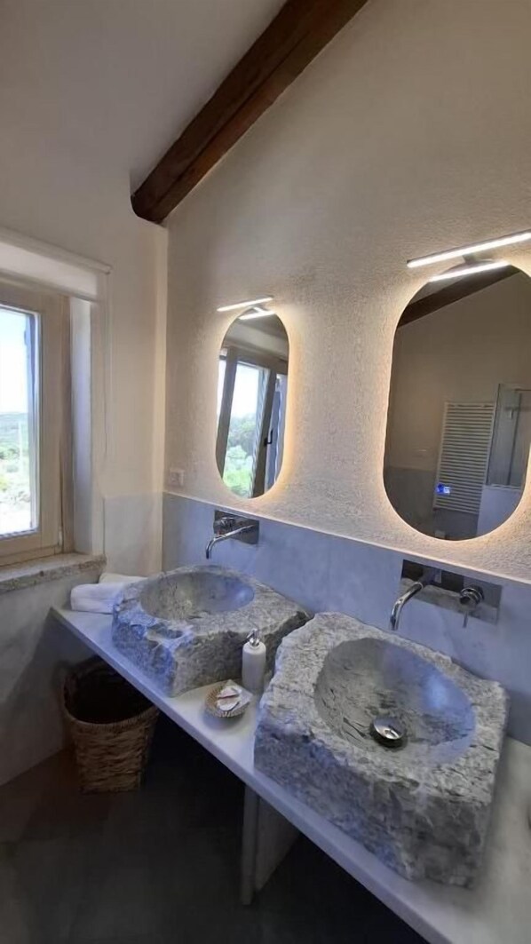 A bathroom with two stone sinks, a window, and a mirror with integrated lighting.