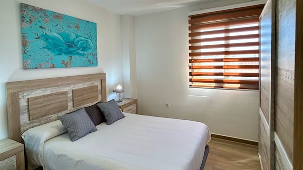 Ein Schlafzimmer mit Bett, Nachttisch, Lampe, einem Fenster mit Jalousien und einer Deckenleuchte.