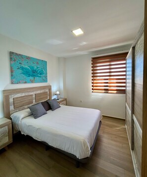 Une chambre à coucher comprenant un lit, une table de chevet, une lampe, une fenêtre avec des stores et un luminaire au plafond.
