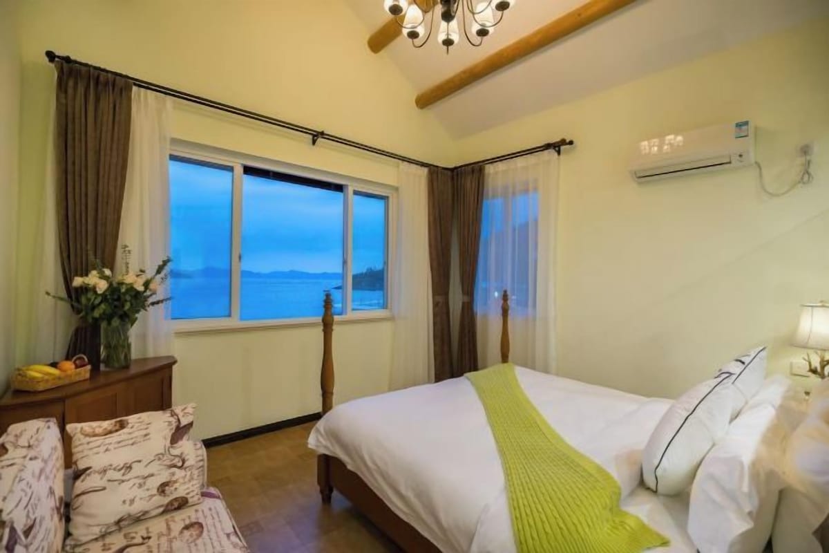 Un dormitorio con una cama, una mesita de noche, una silla, una ventana con cortinas y un equipo de aire acondicionado.