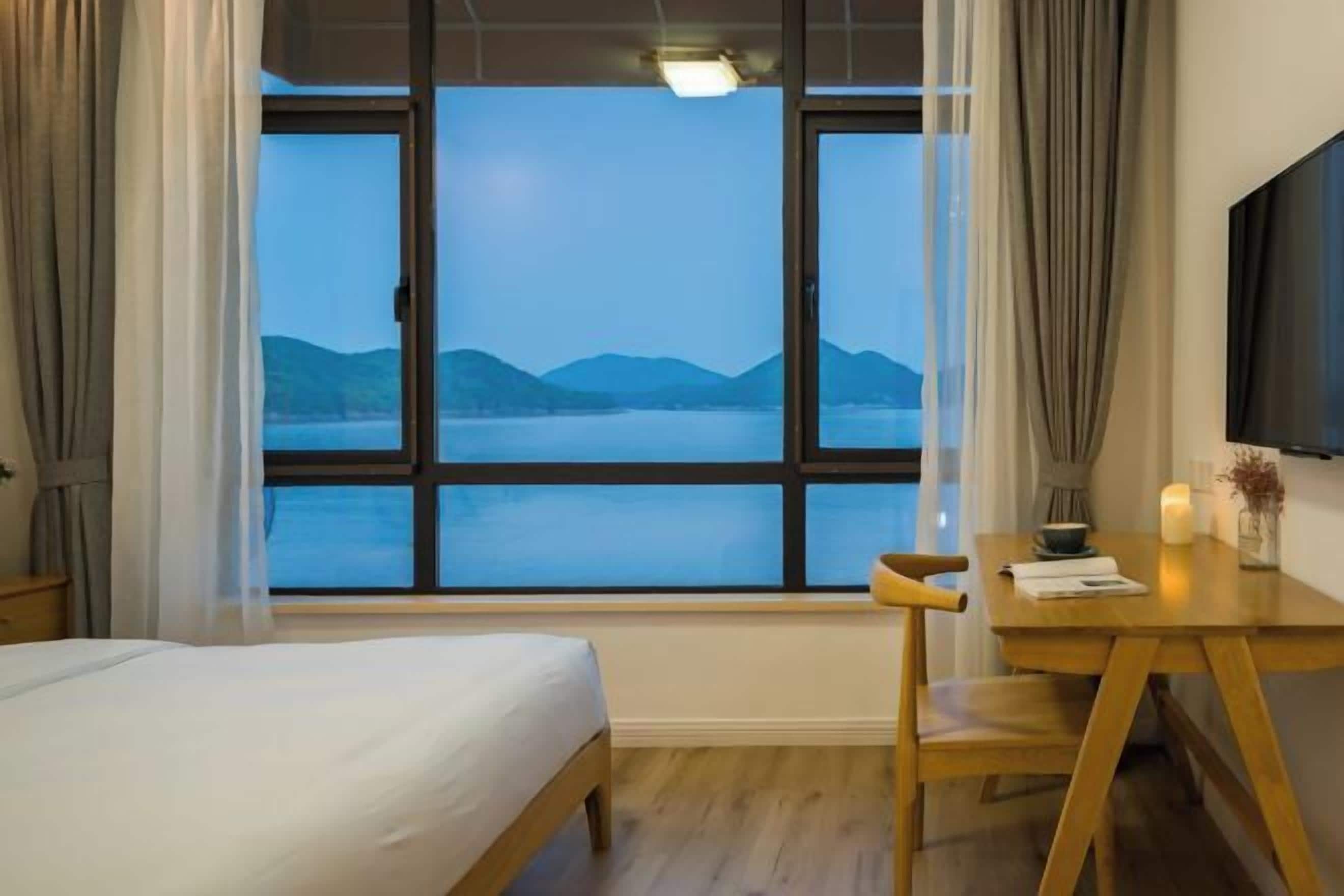Habitació d'hotel amb llit, escriptori de fusta, cadira, televisió i una finestra gran amb vista a una massa d'aigua i muntanyes.