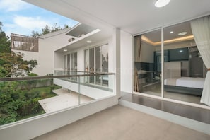 Ein moderner Balkon mit Glasgeländern, Blick auf Grünflächen und ein Einblick in ein Schlafzimmer mit Bett und Vorhängen.