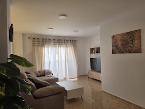 Una sala de estar con un sofá, una mesa de centro, un televisor sobre un mueble bajo, un cuadro en la pared y un ventanal grande con cortinas.