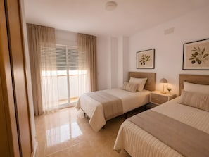 A hotel room with two beds, a large window with blinds, a bedside table with a lamp, and a framed plant on the wall.