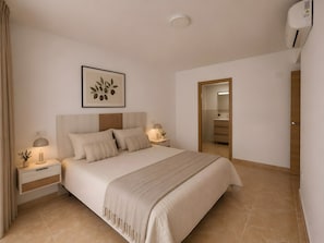 Un dormitorio con cama, mesitas de noche, lámpara de noche, aire acondicionado instalado en la pared y una puerta que da a otra habitación.