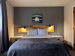 A neatly made bed with a dark textured bedspread, two matching pillows, and a wooden headboard with integrated reading lights. - Alaska Adventure Basecamp • Sleeps 6 • 
• 3BR / 2Ba • Yard + Fire Pit (Anchorage)