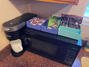 A kitchen area with a coffee maker, a microwave, and a coffee pod holder with various brands.