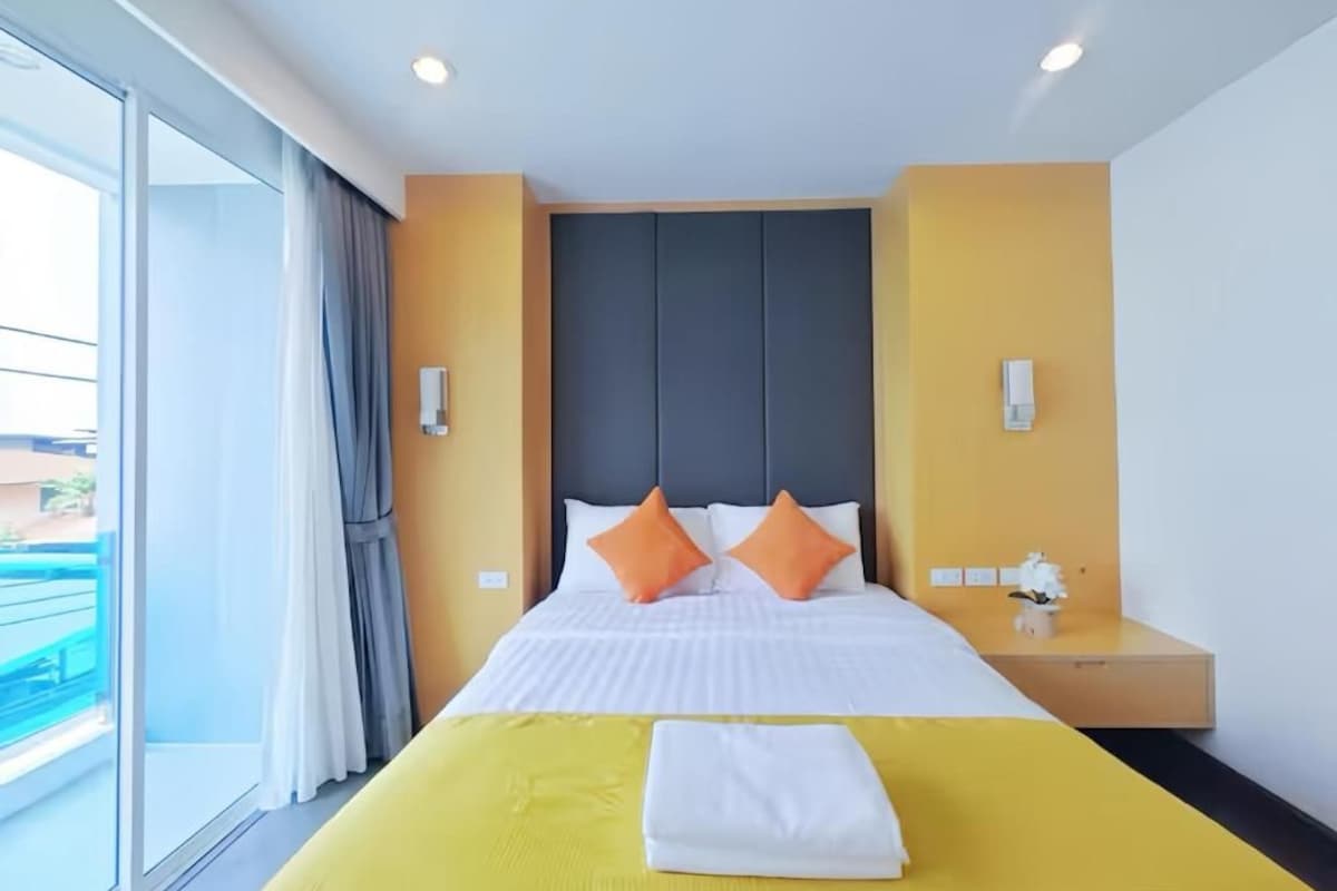 A hotel room with a bed, a nightstand, a window with curtains, and a view of the outside.