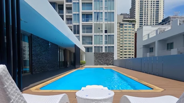 Un espace piscine aménagé sur le toit, avec une piscine à l’eau d’un bleu limpide, des fauteuils de relaxation en osier blanc et un mur en pierre.