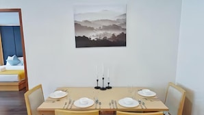 Un coin repas avec une table dressée pour quatre personnes, un tableau accroché au mur et, au fond, un lit avec des oreillers.