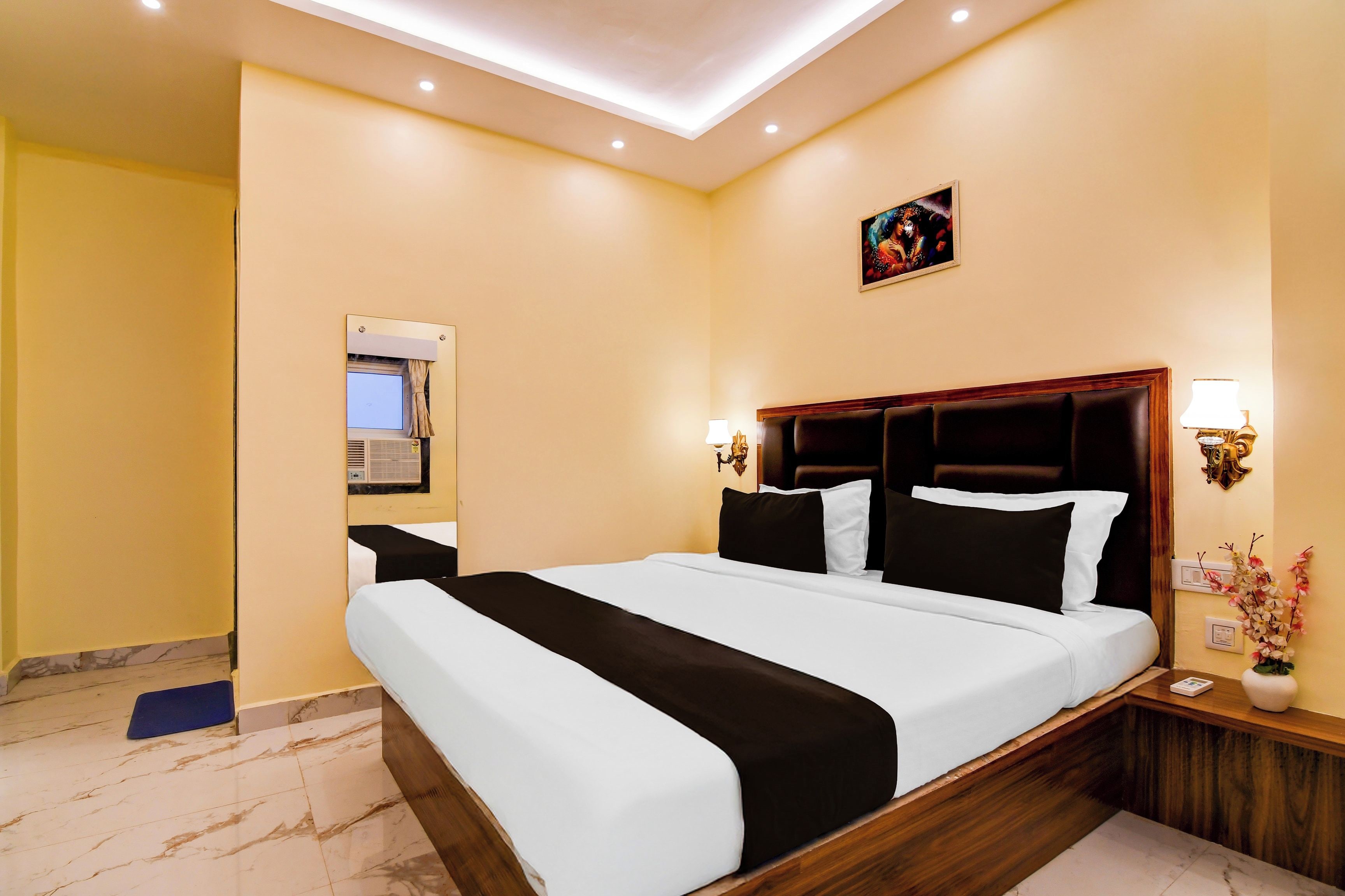 Una habitación de hotel con una cama grande, televisión, aire acondicionado instalado en la pared y una mesita de noche con una lámpara y un florero.