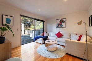 A modern living room with a white sofa, a coffee table, and a floor lamp. There is a large window with a view of trees and a patio outside. - She Oak Treetop Escape (Fairhaven)