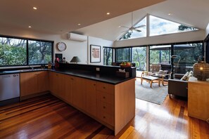 A modern kitchen with wooden cabinets, a black countertop, and a large window overlooking a garden. - She Oak Treetop Escape (Fairhaven)