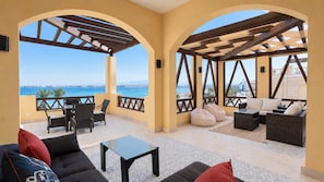 A covered outdoor seating area with a view of the sea, furnished with sofas, chairs, and a table.