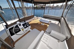 Convés de barco com timão, área de estar acolchoada e vista para a água.