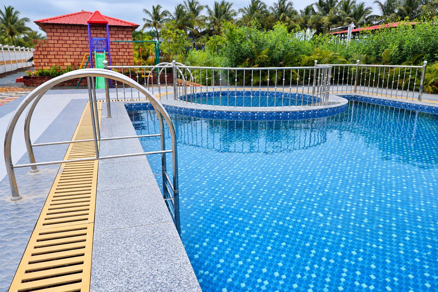 Une piscine dont le bord est recouvert de carreaux, une rambarde de sécurité en métal et, au fond, un petit bâtiment avec un toit rouge.