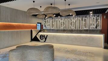 Zona de recepción con diseño moderno, que incluye un mostrador con la palabra 'ey' a la vista, un banco tejido y una pared decorativa con siluetas de árboles.
