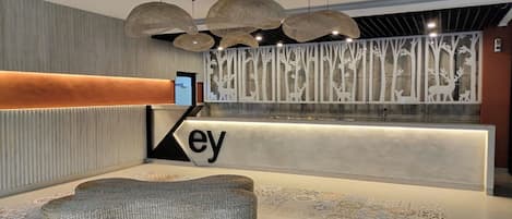 A reception area with a modern design, featuring a reception desk with the word 'ey' visible, a woven bench, and a decorative wall with tree silhouettes.