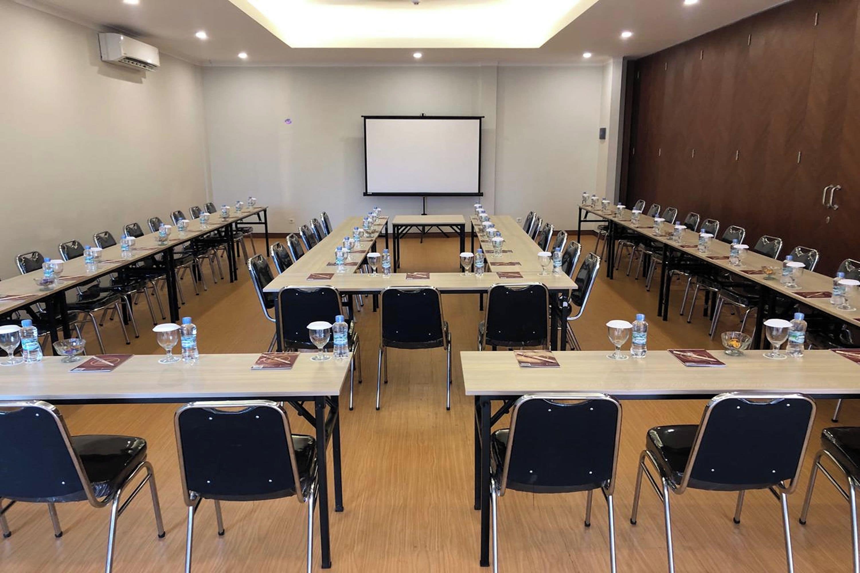 Una sala de conferències amb taules llargues, cadires, una pantalla de projector i una pissarra.