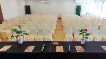 Un salón de conferencias con filas de sillas blancas y una mesa negra con micrófonos y tarjetas de identificación.