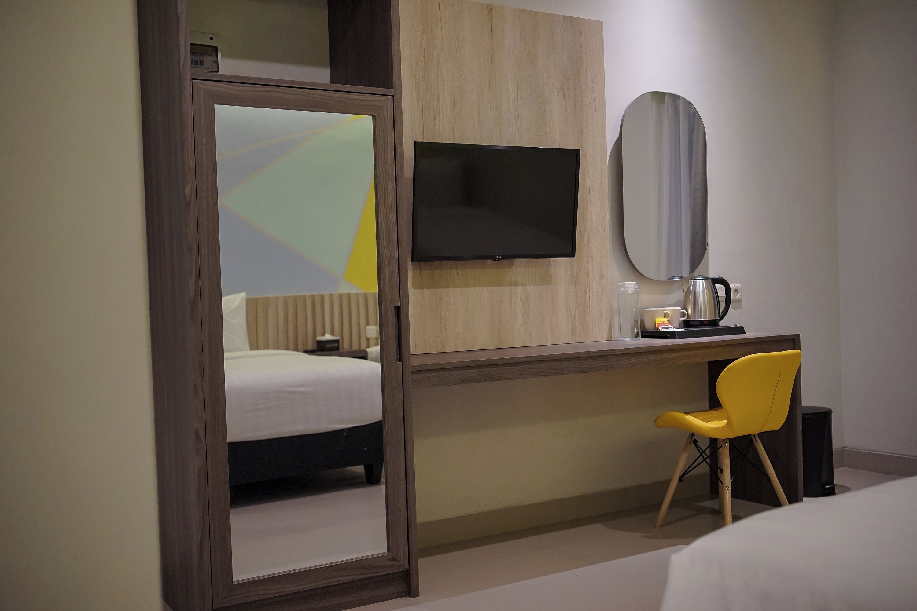Habitación de hotel con una cama, un escritorio con una silla, un televisor de pantalla plana, un espejo y una cafetera.