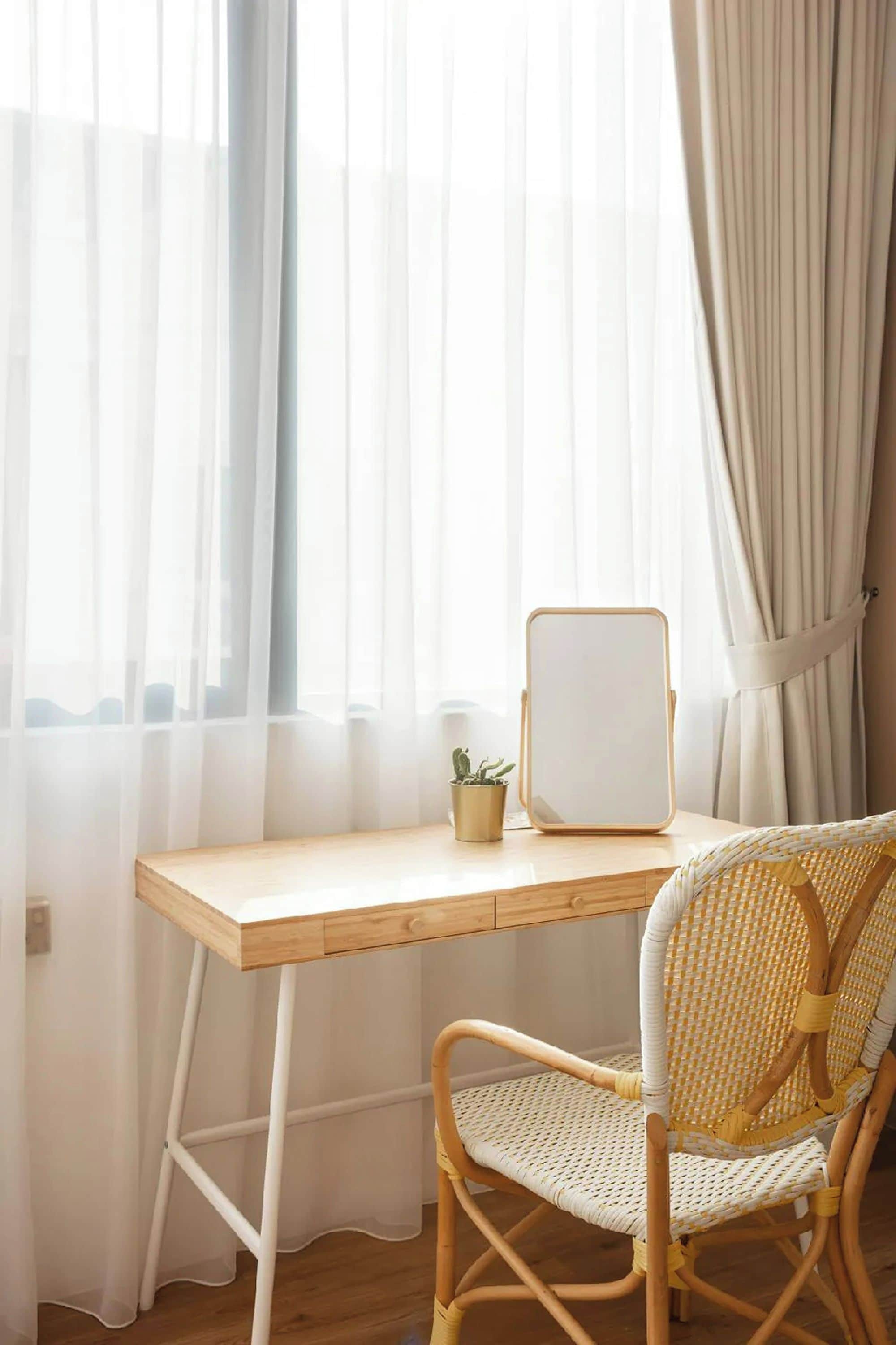 Una habitación con un escritorio de madera, una silla, una pequeña planta en maceta y cortinas traslúcidas.