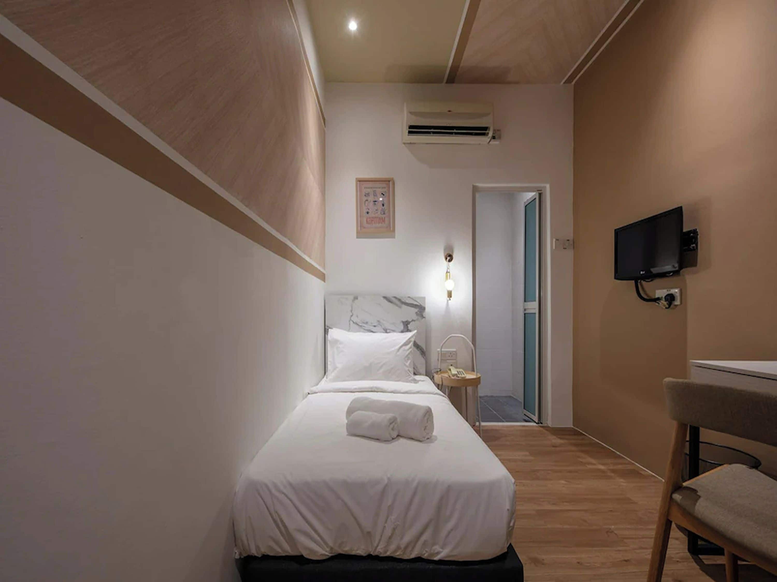 A hotel room with a bed, a desk, a chair, an air conditioning unit, a television, and a door leading to another room.