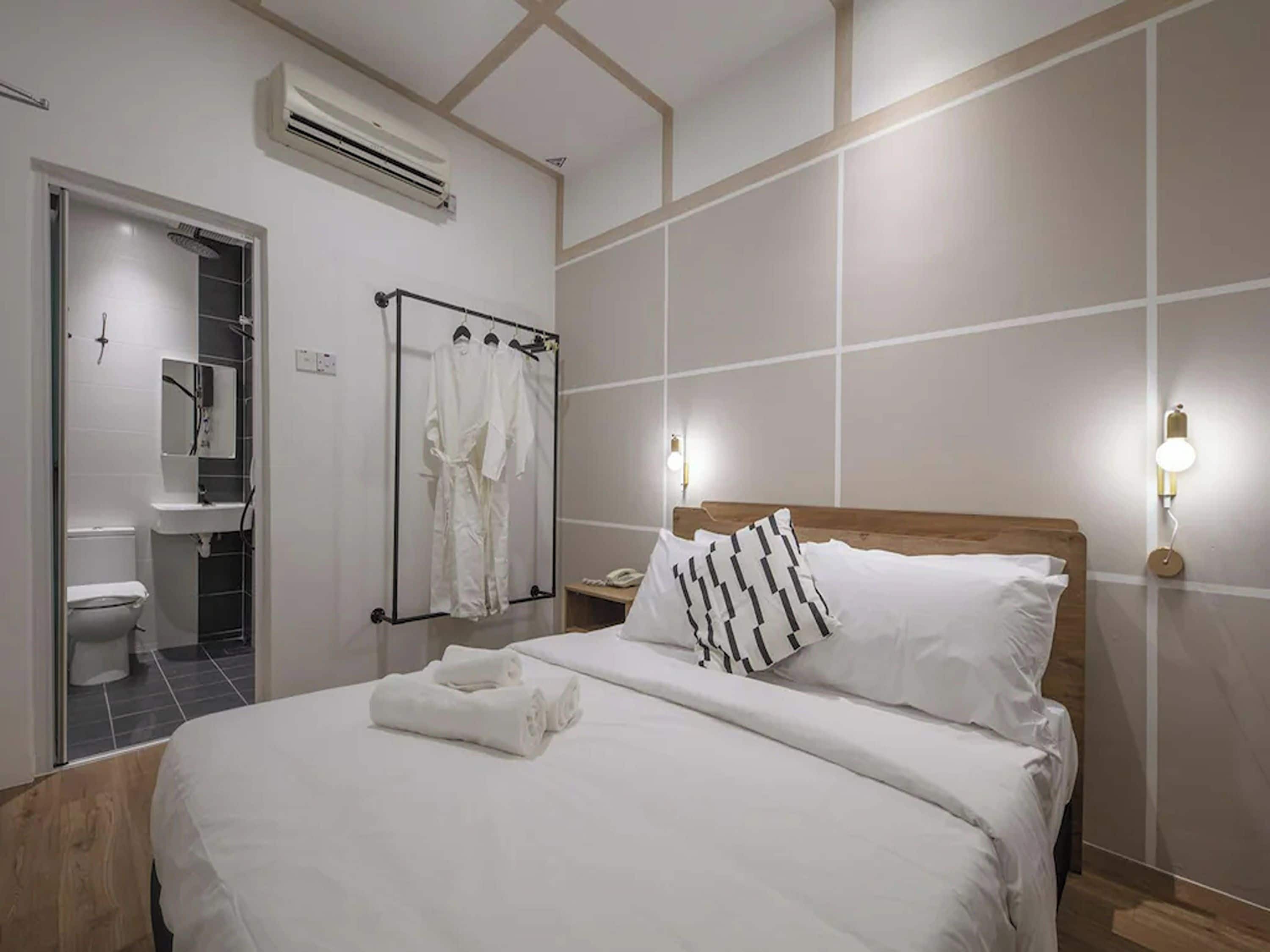 A hotel room with a bed, a bedside table, a nightstand, a wall-mounted light, a towel on the bed, a shower area, and a toilet.