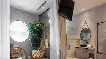 A modern bathroom with a shower, toilet, and sink. There is a large mirror, a television mounted on the wall, and a potted plant.