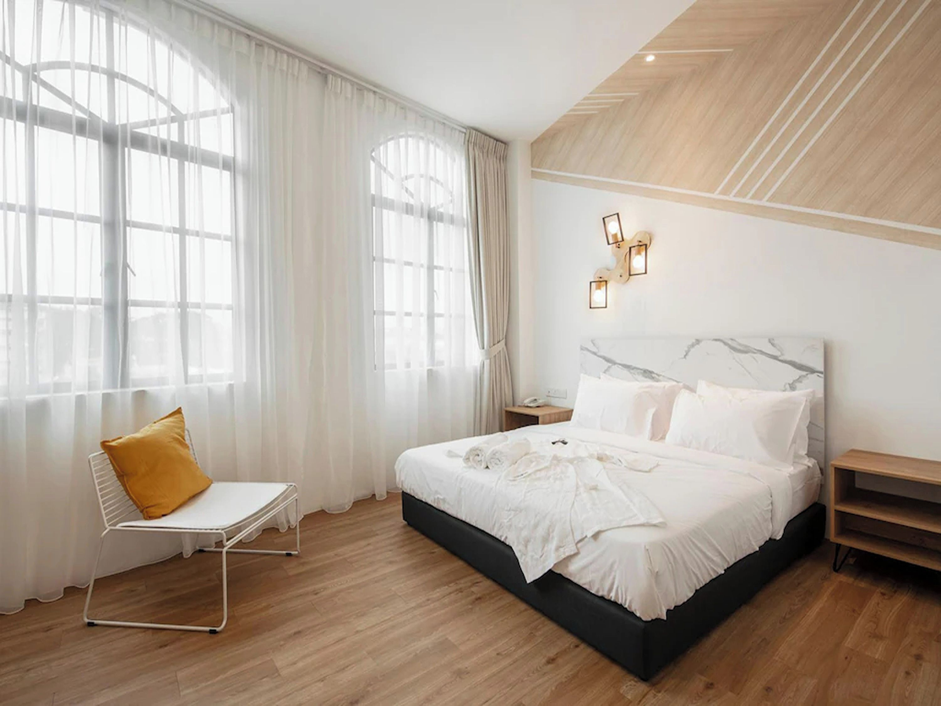 A bedroom with a large bed, a bedside table, a chair, and a window with sheer curtains.