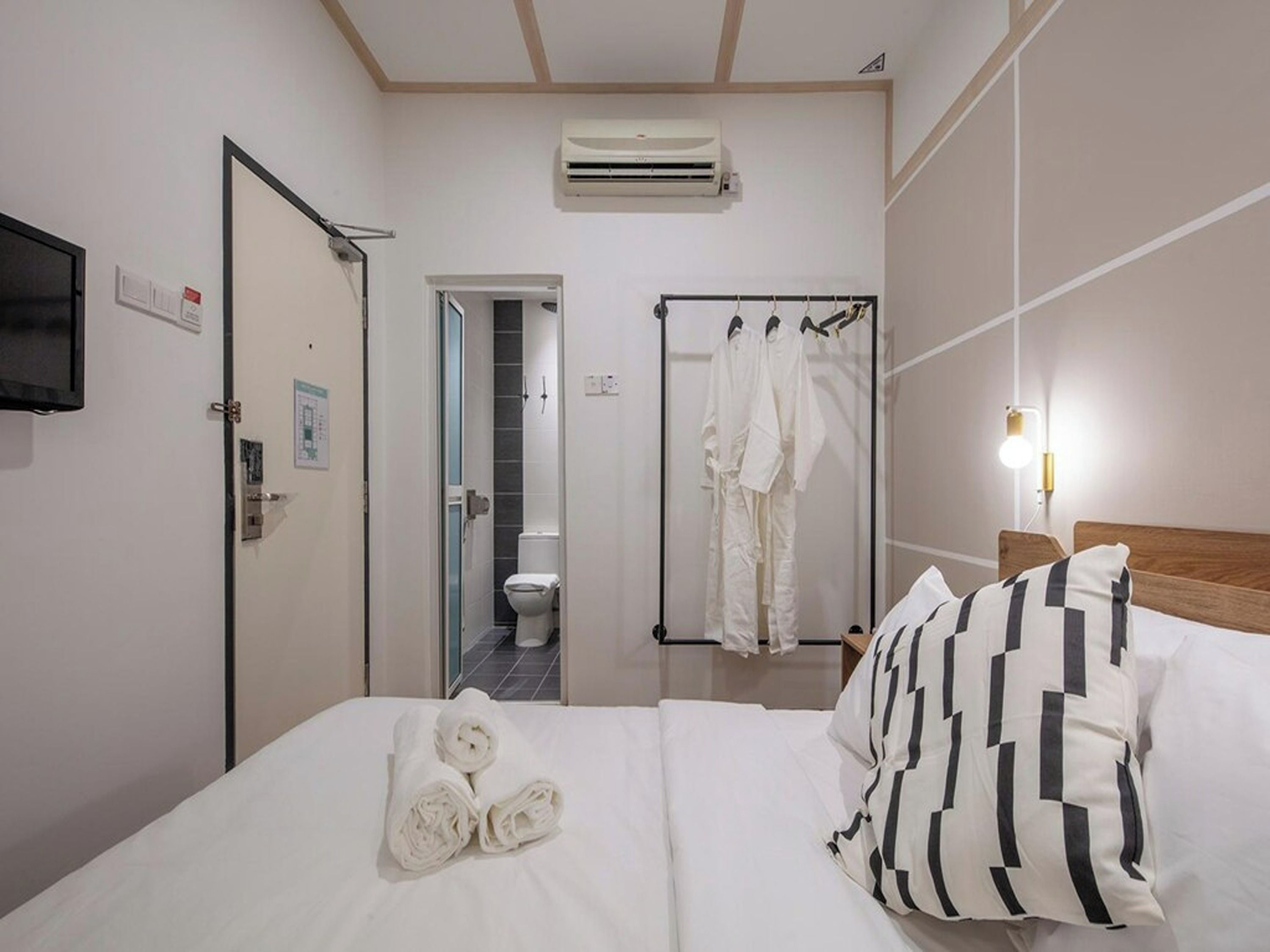 A hotel room with a bed, a TV mounted on the wall, an air conditioning unit, a bathroom with a toilet and shower, and a towel rack with clothes.
