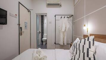 A hotel room with a bed, a TV mounted on the wall, an air conditioning unit, a bathroom with a toilet and shower, and a towel rack with clothes.