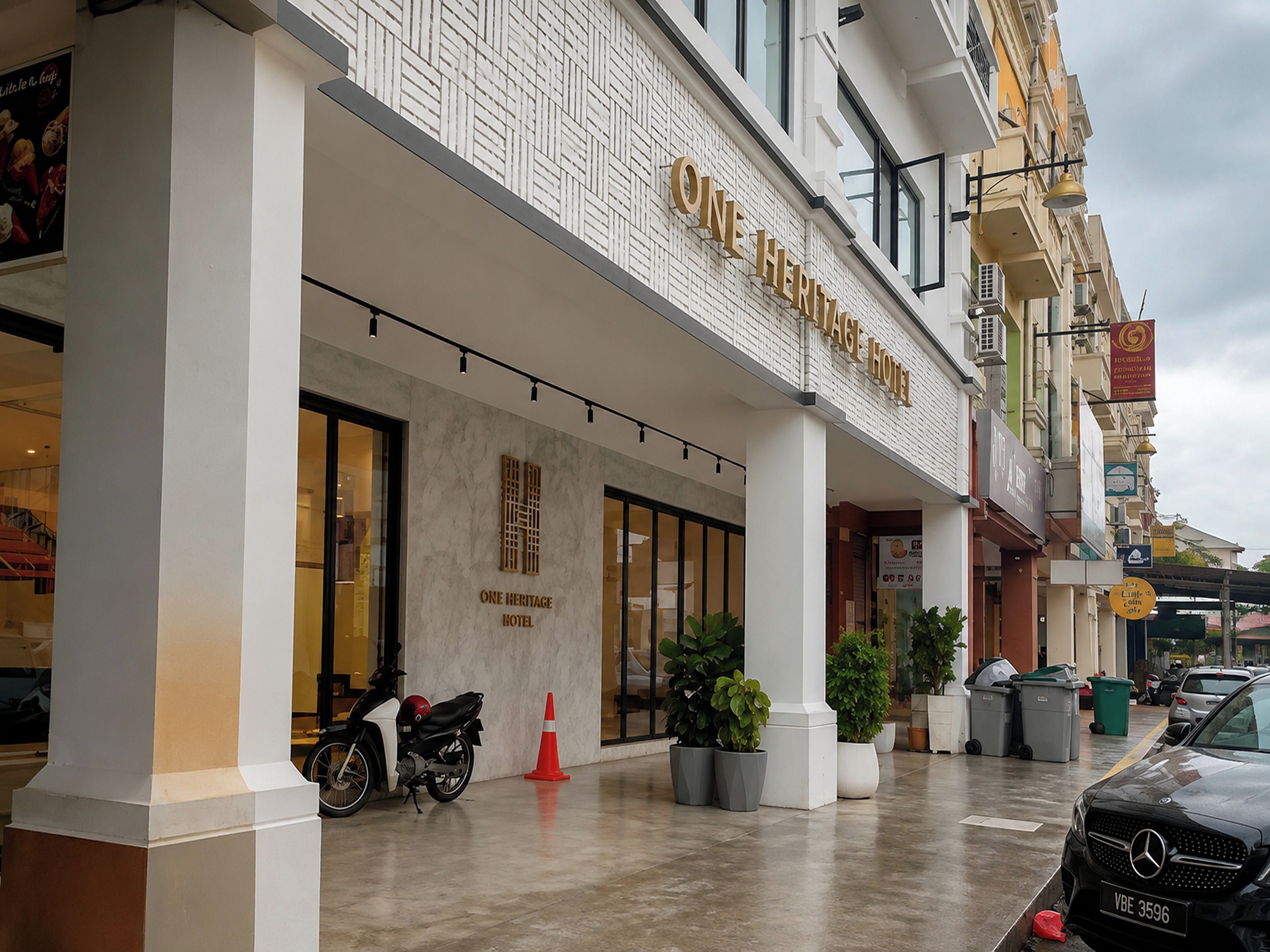 Ein Hotel Eingang mit einem Schild mit dem Schriftzug 'ONE HERITAGE HOTEL', einem Motorrad, das davor geparkt ist, und mehreren Blumentöpfen.