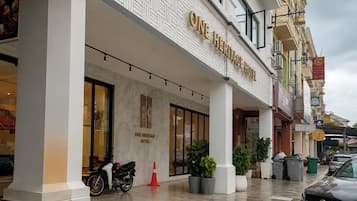 A hotel entrance with a sign reading 'ONE HERITAGE HOTEL,' a motorcycle parked outside, and several potted plants.