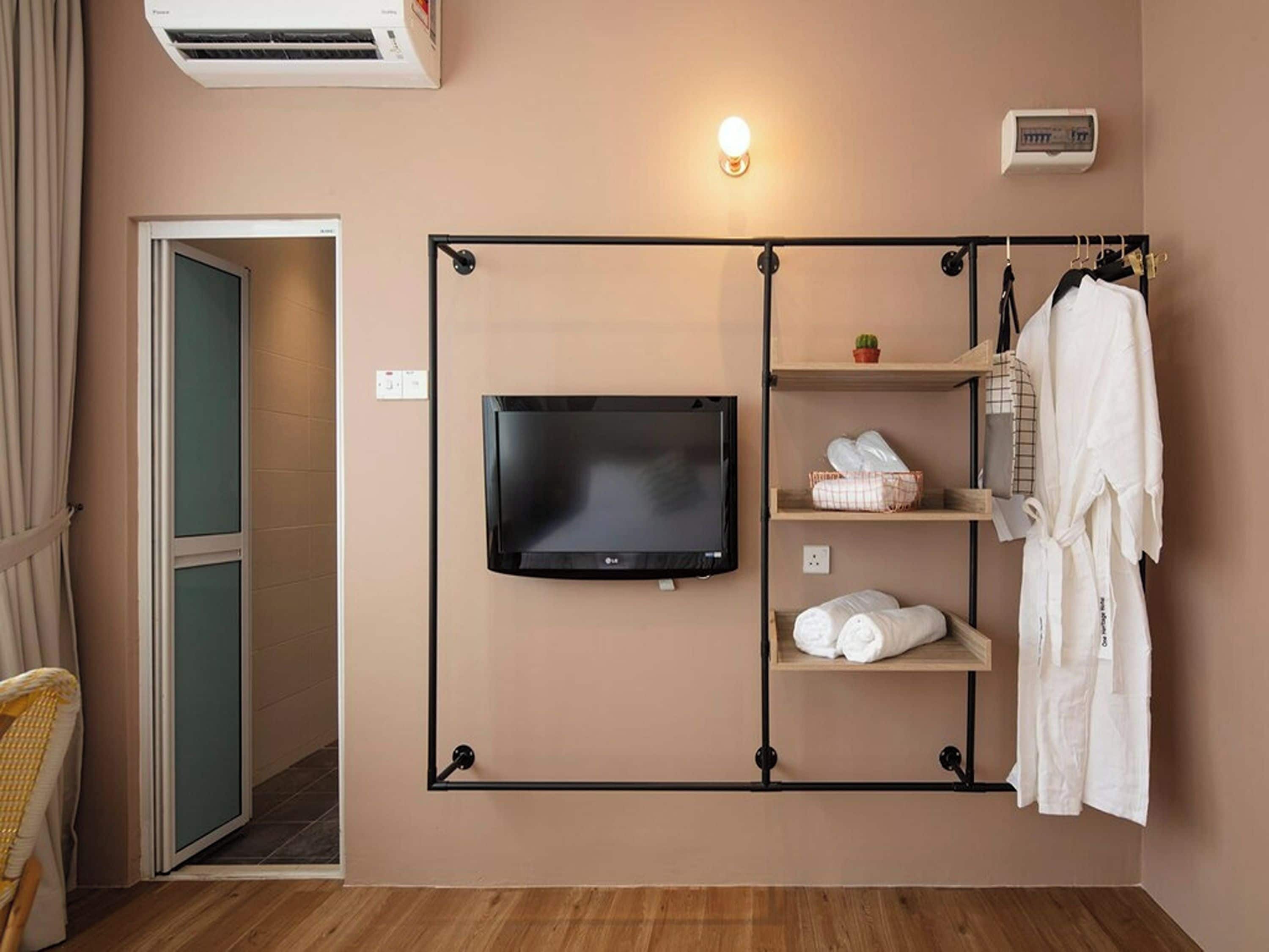 A hotel room with a television mounted on the wall, a robe hanging on a rack, and a shelf with towels and toiletries.
