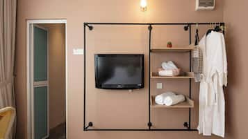 A hotel room with a television mounted on the wall, a robe hanging on a rack, and a shelf with towels and toiletries.