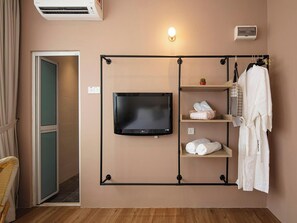 Habitación de hotel con un televisor montado en la pared, una bata colgada en un perchero y un estante con toallas y artículos de aseo.